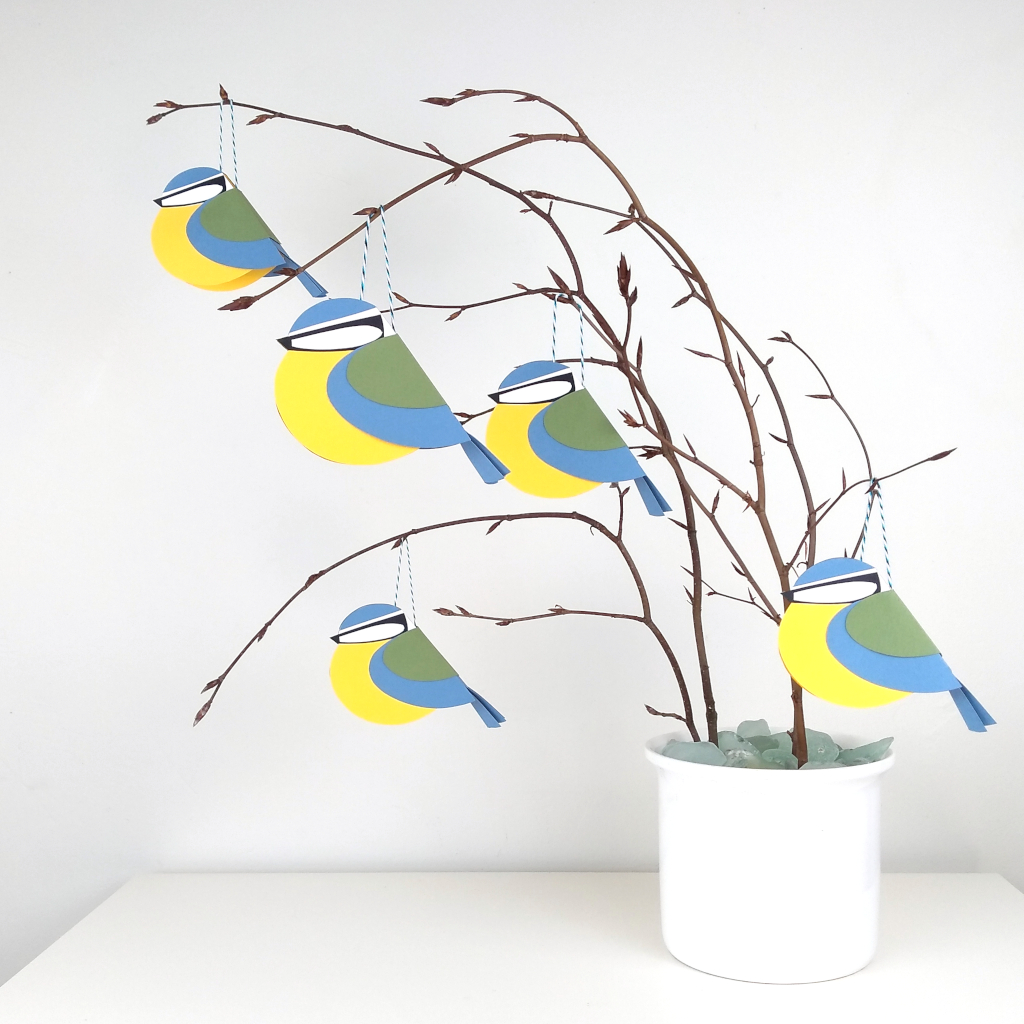 Paper blue tits hanging on twigs in a white plant pot on a white table top against aplain neutral background