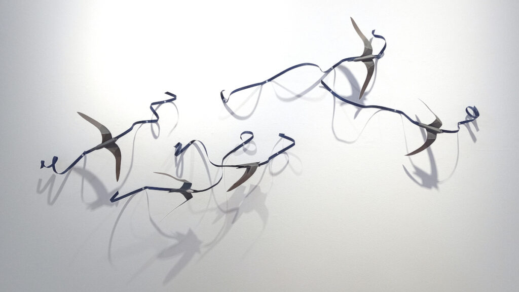 A photograph of a wall intstallation of 5 paper swifts attached to dark blue ribbons of papre that curl and twist in and out from the wall